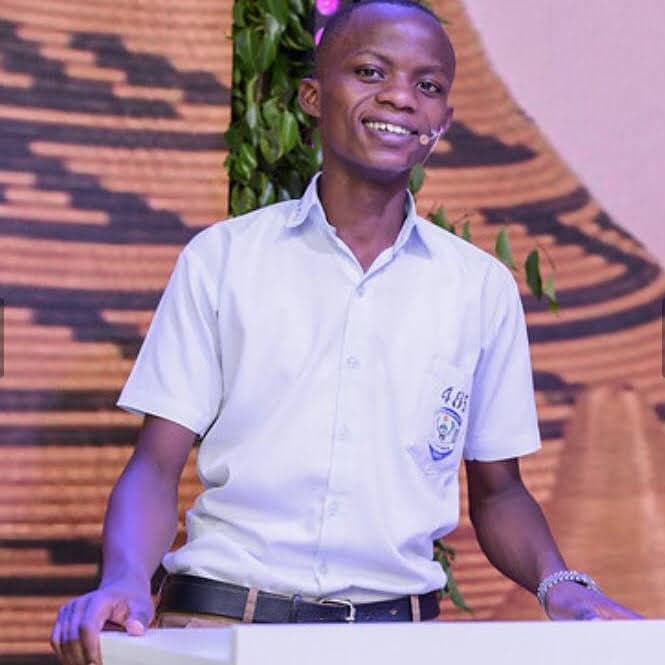 Evode Manirahari at a lectern in school uniform, wearing a headset microphone.