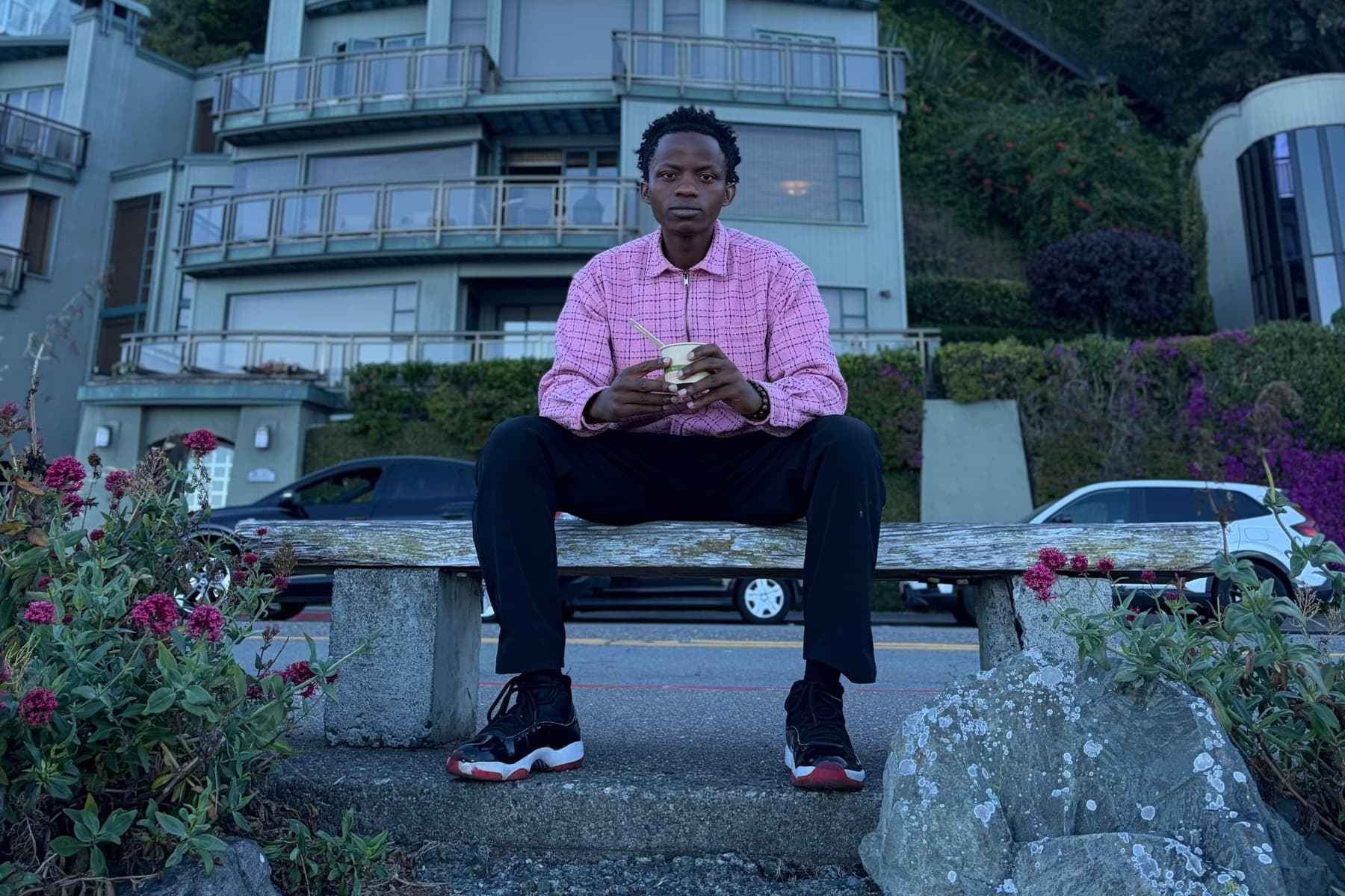 Evode Manirahari seated on a bench beside a road, with hillside houses and trees rising behind him.